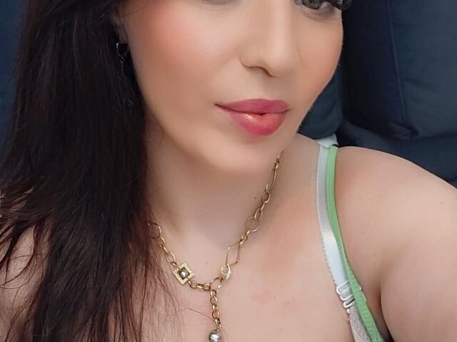 VanessaSquirtt, con labbra rosa, una tracolla verde, una collana a catena dorata e capelli castano scuro, mostra il suo seno carino in un reggiseno verde, sorridendo dolcemente..