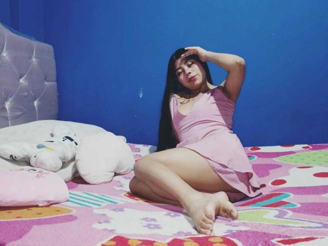 CataMoreno in a pink dress, sitting on a colorful bed with a white stuffed animal, hand in her hair, legs crossed, bare feet, blue wall, long dark hair.
