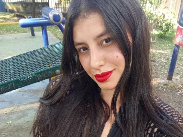 Madisson Fontaine, avec ses longs cheveux noirs, son rouge à lèvres rouge et une petite tache de rousseur sur sa joue droite, souriant à l'appareil photo, portant un haut en maille noire, dans un parc..