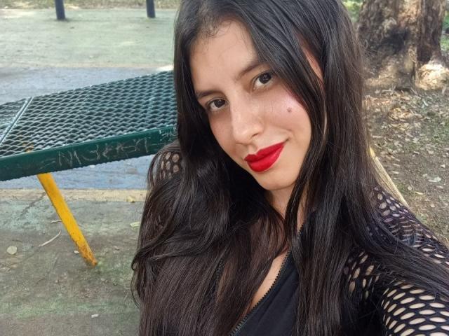 MadissonFontaine with long black hair, red lipstick, and a black fishnet top, showing off her tits in a selfie outdoors near a green metal table.