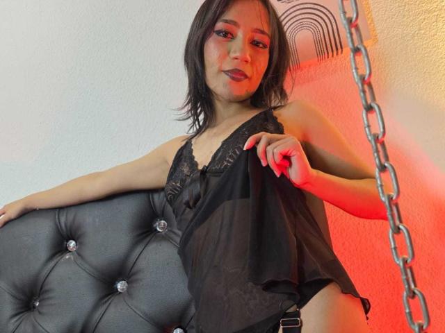 HenutmireRossi with black lace lingerie, holding up a sheer black top, sitting on a tufted gray chair, red and orange lighting, and a chain in the background.