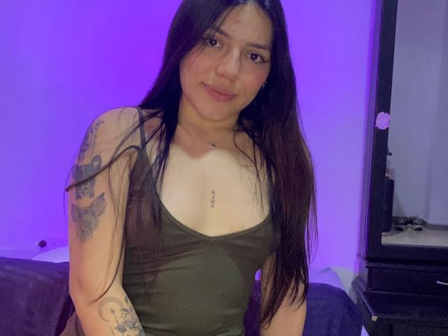 SophiaMarx has long black hair, a green tank top, small tits, a tattoo on her right arm, and a nipple piercing, smiling with purple lighting in the background.