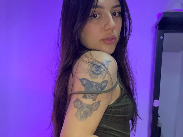 SophiaMarx with long dark hair, green tank top, and tattoos of bats on her arm, looking over her shoulder at the camera. Purple lighting. Soft expression.