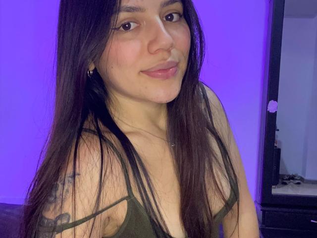 SophiaMarx with long dark hair, green top, small tits, tattoo on left shoulder, subtle smile, purple background, mirror on right, casual selfie.