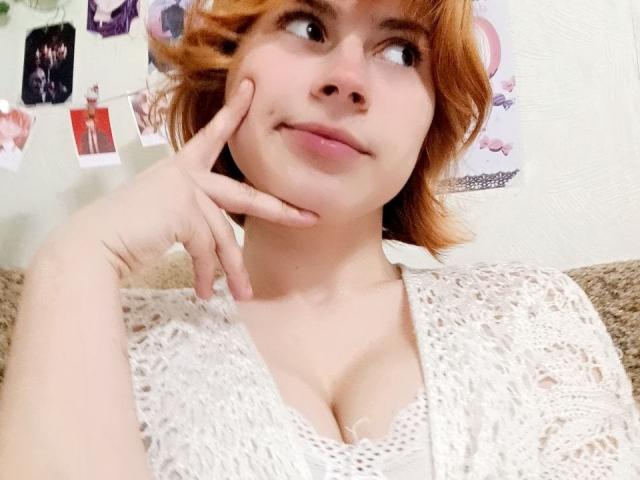 MireilleSpotwood with short red hair, wearing a white lace top, has her chin in her hand, looking to the side with a slight smile, showing her tits.