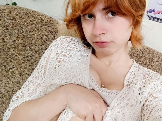 MireilleSpotwood with red hair, wearing a white crocheted top, arms crossed, showing small tits, sitting on a brown textured couch, looking slightly to the side.