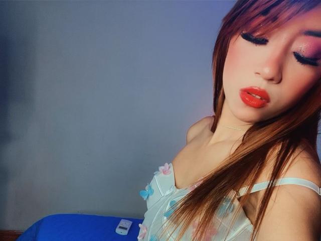 KarlyxRose tiene el cabello largo y castaño, lápiz labial rojo y pestañas negras y gruesas, y lleva un vestido floral azul claro con botones pastel, y un cojín azul en primer plano..