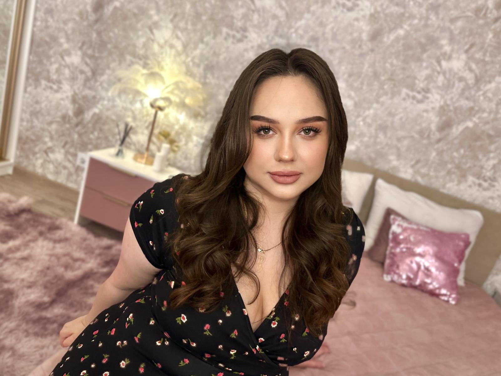 MaddyCroft with wavy brown hair, wearing a black floral dress, has her tits slightly visible, sitting on a pink bed, looking at the camera with a slight smile.