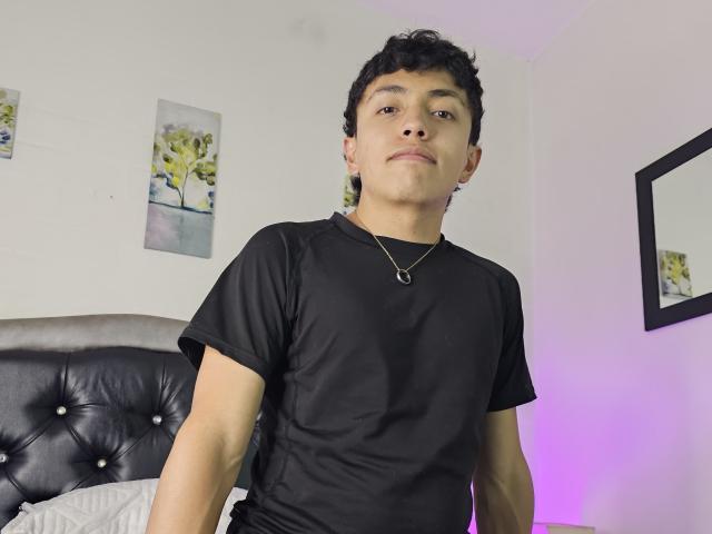 MaliciusKing, vestindo uma camiseta preta, tem cabelo curto e cacheado, de cor preta, e um pequeno piercing no nariz. Ele está em um quarto com uma cabeceira preta estofada e obras de arte nas paredes..