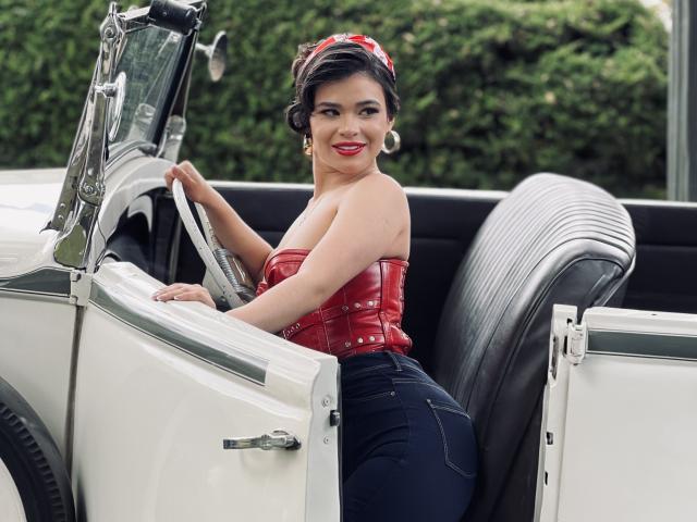 PaulinaValley, vêtue d'un bustier en cuir rouge, montrant ses fesses, assise dans une voiture vintage blanche, cheveux foncés attachés avec un bandeau rouge, souriante, portant un jean bleu et de grandes boucles d'oreilles..