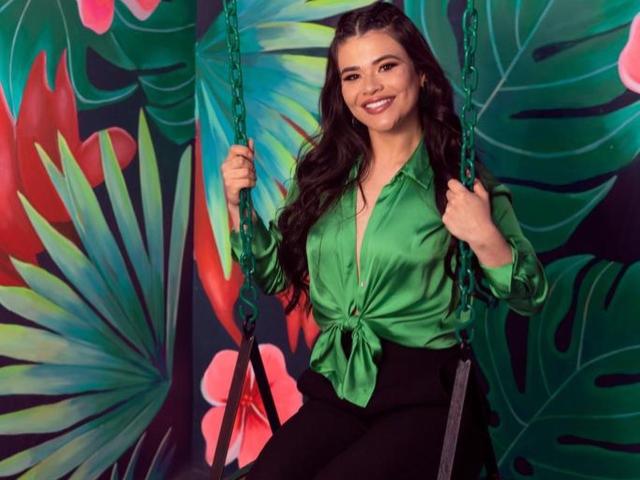 PaulinaValley met lang donker haar, een groene satijnen blouse die aan de voorkant is dichtgeknoopt, zwarte broek, zittend op een groene schommel, lachend, met de kettingen van de schommel in haar handen, achtergrond met een tropisch bladpatroon..