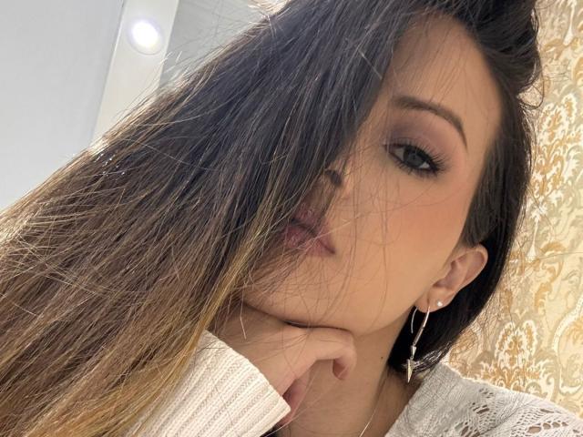 HannaRobertss has long brown hair covering her right eye, wearing a white lace top, with a hand under her chin, and a silver cross earring in a selfie.
