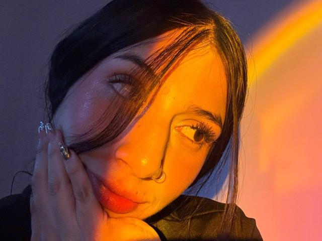 O rosto de Sophie Beaumont, com cabelo preto, piercing no nariz e pele brilhante, iluminado por luzes laranja e amarela, com uma mão adornada com um anel tocando sua bochecha..