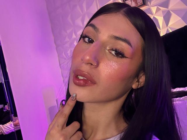 Sophie Beaumont tiene una piel brillante con un tono rosado, cabello largo y oscuro, cejas gruesas, párpados brillantes y labios rojos y brillantes, con un dedo tocando su barbilla. Hay una luz púrpura en el fondo..