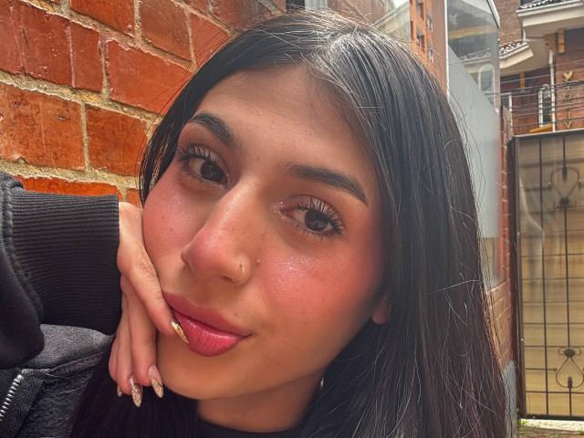 Het gezicht van Sophie Beaumont, met lang zwart haar, roze lippen en glanzende ogen, leunt met haar kin op haar hand, waarbij haar verzorgde nagels te zien zijn, voor een muur van rode bakstenen..
