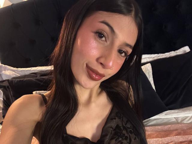Sophie Beaumont heeft glanzende, roze wangen, lang zwart haar en een schattige glimlach. Ze draagt een zwarte top van kant, waardoor haar kleine borsten en een lichte decolleté zichtbaar zijn..