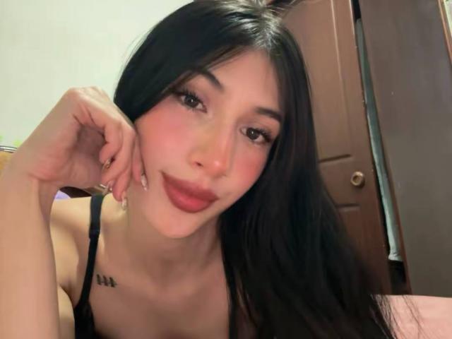 Sophie Beaumont, com cabelos pretos longos, olhos castanhos e batom rosa, vestindo uma blusa preta, mostrando uma tatuagem no peito, com o queixo apoiado na mão..
