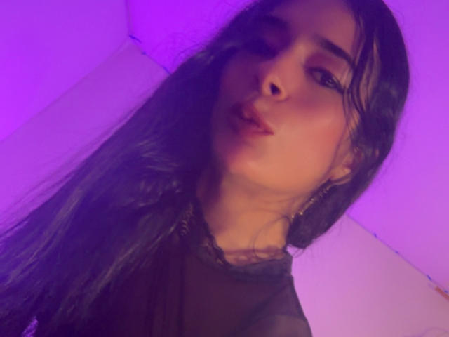 ArielNitzche has long black hair, dark eyebrows, and full lips with a slight pout. She's wearing a sheer black top, and the purple light highlights her skin.