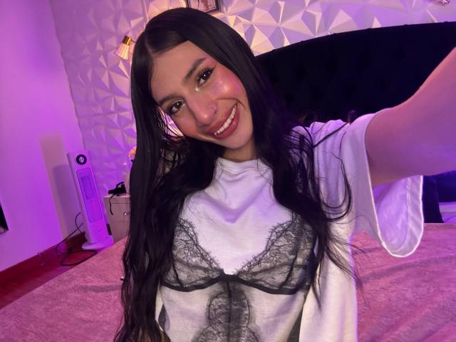 SophieBeaumont with long black hair, smiling, wearing a white shirt with black lace bra, holding out arm for selfie, purple lighting, white wall, black headboard, standing in bedroom.