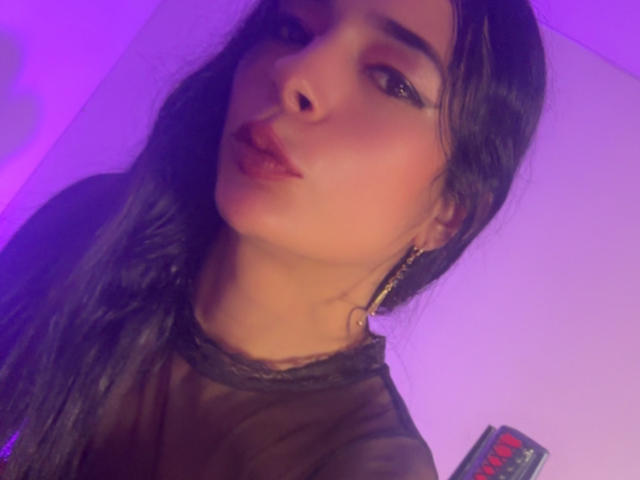 ArielNitzche with pouty lips, dark wavy hair, and hoop earrings, wearing a sheer black top, lit by purple light. Her brown eyes and full lips are prominent.