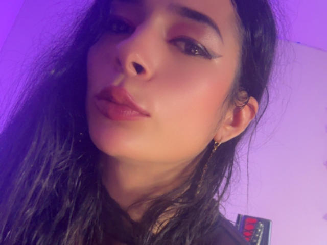 ArielNitzche has long dark hair, full pink lips, and subtle eyeliner with winged edges. She's looking at the camera with a slight, confident smile. Purple light highlights her face.
