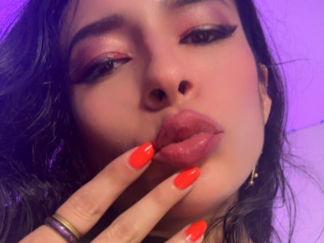 ArielNitzche's face with pink eyeshadow, red lipstick, and shiny red nails gently touching her lips. She has dark hair and a purple light background.