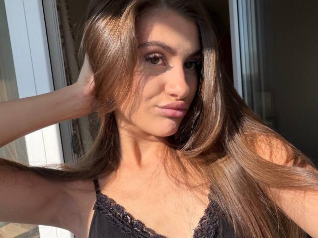 EllenKienbaum has long, straight brown hair, a cute face, and is wearing a black lace top. She's touching her hair with a relaxed, confident expression.