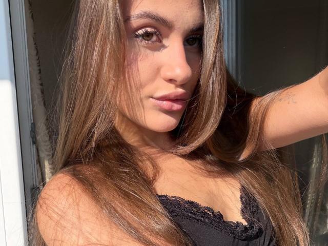 EllenKienbaum has long brown hair, light brown eyes, and is wearing a black lace top, showing her small tits and a tattoo on her right arm, in sunlight.