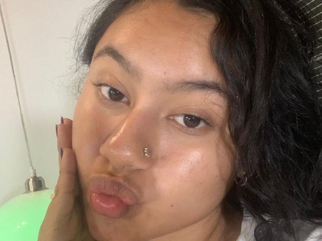 NoiraVale's face with dark curly hair, brown eyes, and a nose piercing. She's puckering her lips, with a hand touching her cheek. Her skin is shiny and she's looking at the camera.