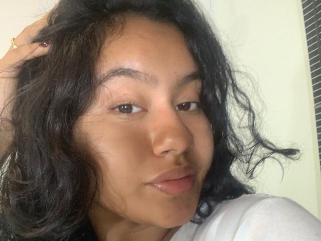NoiraVale with wavy black hair, brown eyes, and light brown skin, slightly smiling, touching her head with a hand, wearing a white shirt, no visible genitals or breasts.