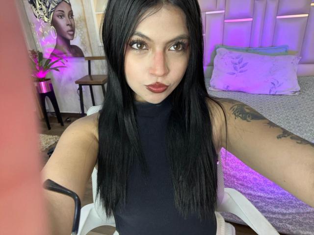 NyraVanel with long black hair, dark eyes, and red lipstick, wearing a black sleeveless top, showing a tattoo on her right arm, taking a selfie in a bedroom.