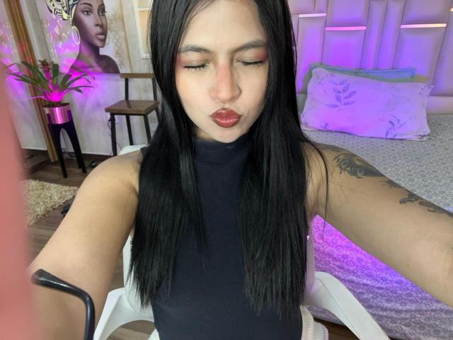 NyraVanel with long black hair, red lipstick, closed eyes, black sleeveless top, tattoo on right arm, sitting on white chair, purple and pink bedroom lighting in background.