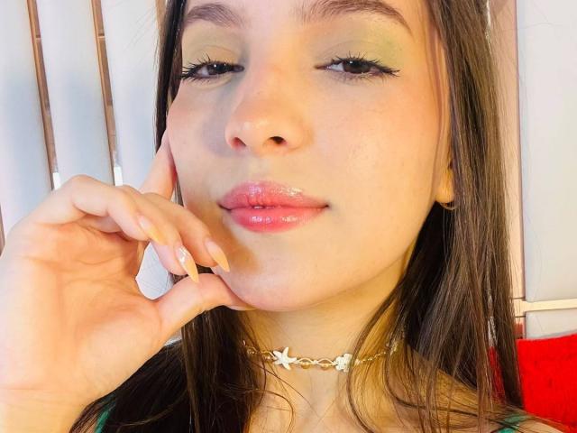 KatarinaNovak has glossy pink lips, green eyeshadow, long brown hair, and a gold chain necklace. She's touching her cheek with manicured nails, smiling slightly.