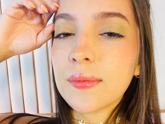 KatarinaNovak with glossy pink lips, green eyeshadow, long black eyelashes, shiny nose, and brown hair, touching her eyebrow with a hand, wearing a gold necklace.