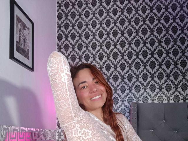 MadisonKoper, avec des cheveux bruns, souriante, portant un haut blanc en dentelle à manches longues, bras levé, montrant ses seins, assise sur un lit gris capitonné, dans une pièce à motifs..