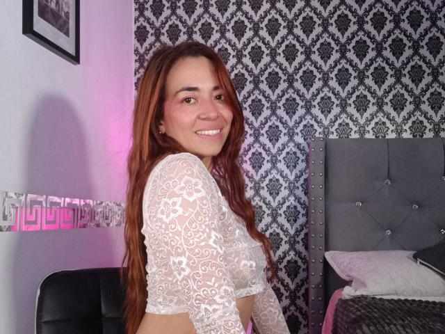 MadisonKoper with long red hair, smiling, wearing a white lace crop top, showing her tits, in a bedroom with a grey upholstered headboard and black patterned wallpaper.