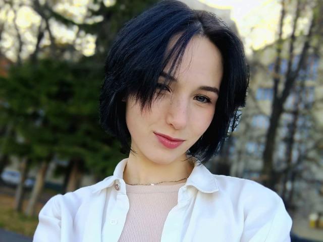 Nathalie Dutt tem cabelo preto curto, pele clara e um nariz pequeno, com batom rosa. Ela está vestindo uma camisa branca por cima de uma blusa rosa texturizada, e está sorrindo discretamente..