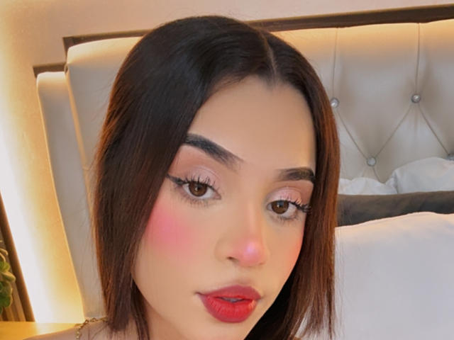 LunaVali has dark brown hair, pink blush, red lipstick, and long eyelashes, with a slightly upturned nose and brown eyes. She's sitting against a tufted headboard.