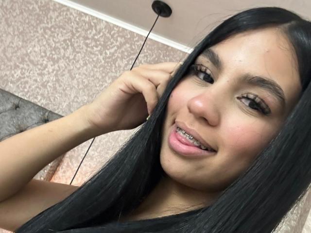 DalilaTennesse with long black hair, slightly parted lips showing metal braces, brown eyes, and a hand in her hair, smiling softly.