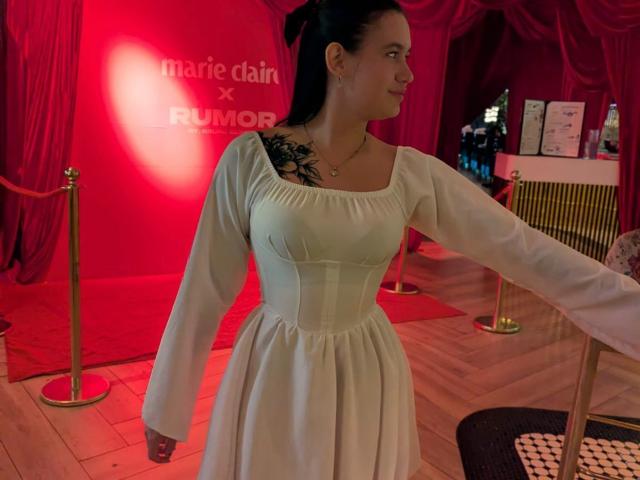 Samantha Juyce, com os seios levemente visíveis sob um vestido branco, tem uma tatuagem preta no ombro direito, cabelo escuro e está estendendo o braço, em pé em um quarto com iluminação vermelha..