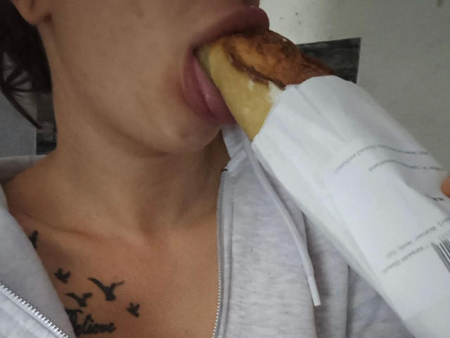 Leonora, con un panino ripieno di salsiccia in bocca, indossa una felpa grigia, ha un tatuaggio di uccelli sul petto, ha un foglio di carta bianco che avvolge il panino, è visibile la clavicola e si vede solo una parte del viso..