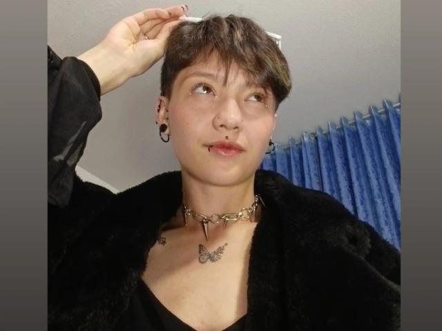 LissyLuxe met kort bruin haar, een zwarte bontjas, een gouden ketting, een vlindertatoeage en zwarte oorbellen, kijkt opzij met een lichte glimlach..