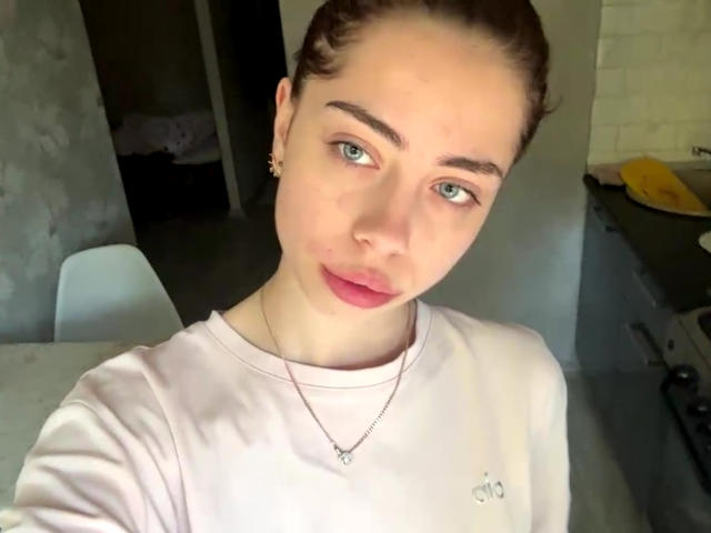 AureaVandale with light skin, blue eyes, and pink lipstick, wearing a pink shirt, small necklace, and earrings, has brown hair pulled back, looking slightly to the side.