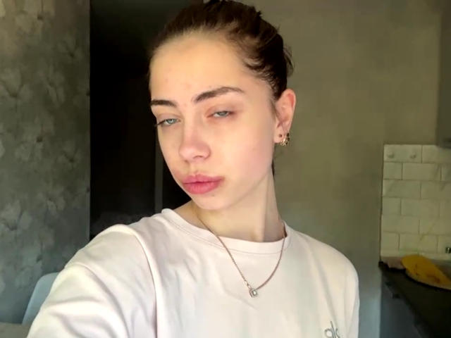 AureaVandale with pouty pink lips, light skin, green eyes, brown hair in a bun, wearing a white shirt and gold necklace, looking slightly to the side.