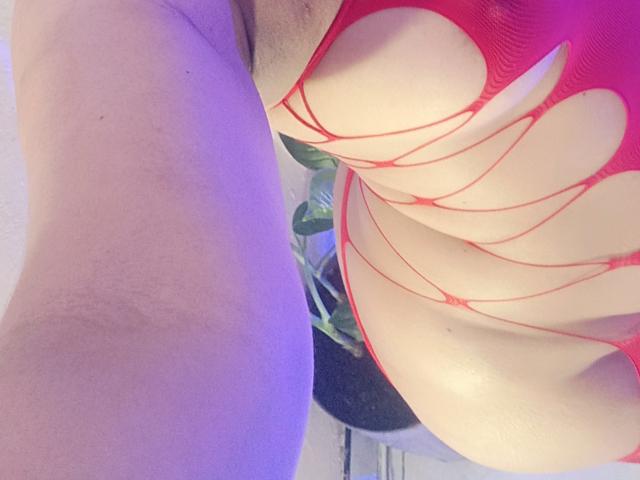 FetishLoverAna's red, cut-out, net-like lingerie highlights her curvy, pale-skinned ass and large tits, with visible lines of red fabric and a green plant in the background.