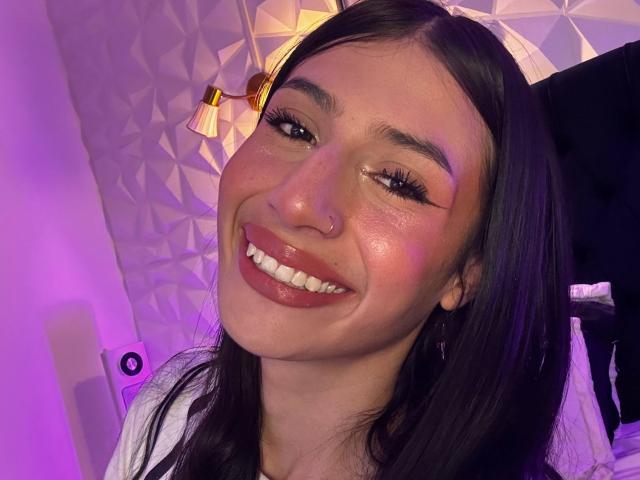 Sophie Beaumont mit einem glänzenden, rosa schimmernden Gesicht, glänzlichen Lippen, einem Lächeln, dunklem, glattem Haar, einem Nasenring und langen Wimpern, in einem Selfie mit violettem Licht, das ihr weißes Hemd und den schwarzen Stuhl zeigt..