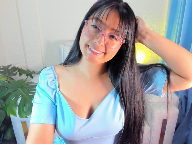 SofiiaThooner with long black hair, pink glasses, and a light blue top showing her tits, smiling, hand in hair, green plant in background, soft light.