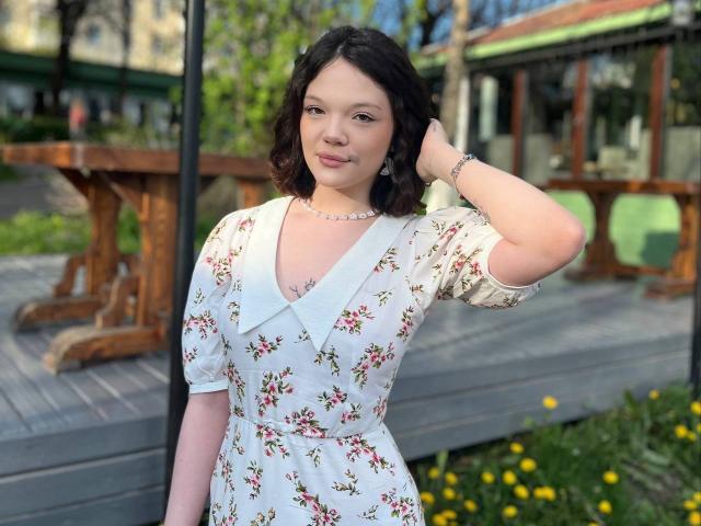 Tawana Shillinger mit kurzen, schwarzen Haaren, die ein Blumenkleid, eine weiße Bluse und eine Perlenkette trägt, mit einer Hand im Haar, steht neben einem Holztisch, im Vordergrund befinden sich grüne und gelbe Blumen..