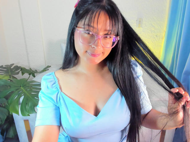 SofiiaThooner with long black hair, pink glasses, blue dress, holding hair, smiling, light skin, medium tits, pink hairclip, green plant, yellow and blue wall in background.