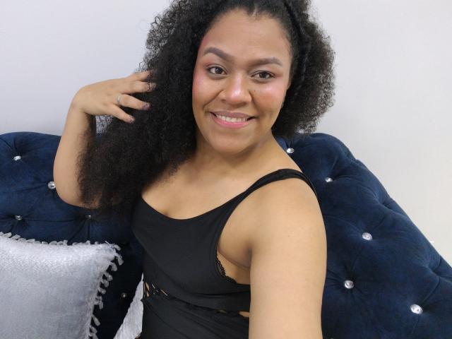 KloheEvans with curly black hair, smiling, wearing a black dress, sitting on a blue tufted couch, holding her hair. She has pink blush on her cheeks, cute.